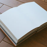 An open blank notebook with white pages lies on a brown wooden floor.