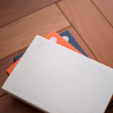 A stack of minimalist hardcover notebooks with circular cutouts in white, orange, and navy blue, arranged diagonally on a warm brown wooden surface.