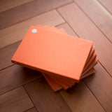 A stack of orange hardcover notebooks with a small white circular dot on the top cover, placed on a wooden floor.