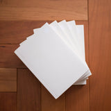 A stack of plain white rectangular notebooks or booklets fanned out on a warm brown wooden floor.