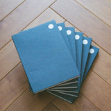 A stack of five textured blue notebooks with white circular page markers, arranged in a fan shape on a light brown wooden floor.