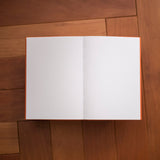 An open blank notebook lying flat with white pages and a thin orange border, placed on a brown wooden floor.
