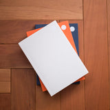 Three hardcover notebooks stacked on a wooden floor, with the top notebook in white, the middle in orange, and the bottom in navy blue.